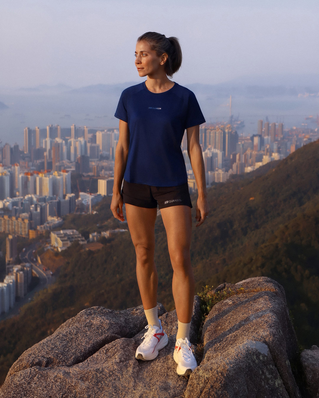 Female trail runner wearing ANTA GUANJUN 100% Merino wool trail running T-shirt and shorts, standing on a mountain ridge overlooking Hong Kong at sunrise